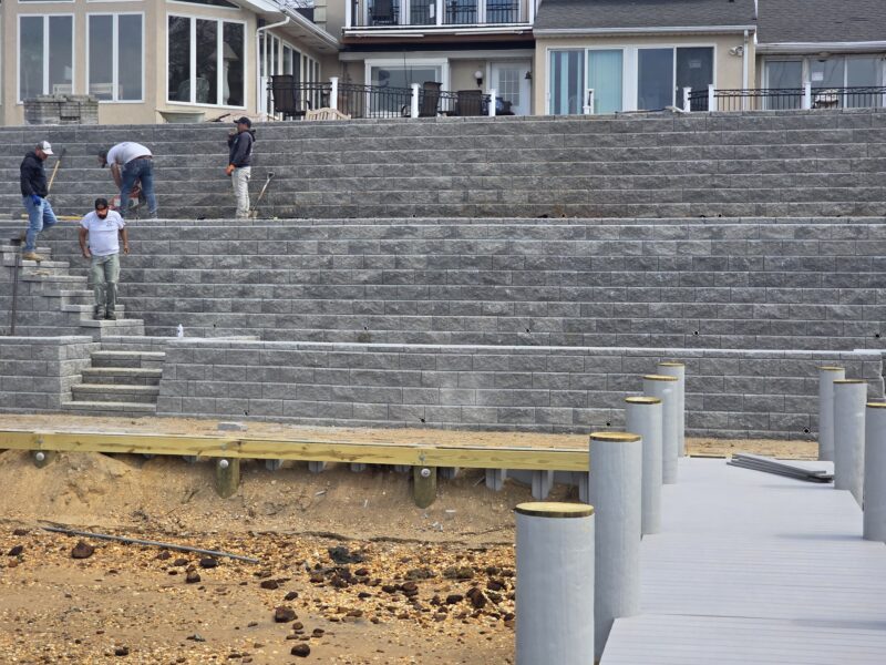 Construction workers standing on a tiered gray stone wall beside a waterfront, with houses in the background and a wooden platform nearby.