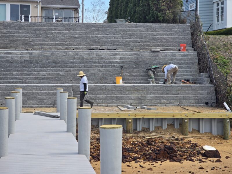 Construction workers build a gray stacked concrete retaining wall behind a yard, with dirt and tools in the foreground.