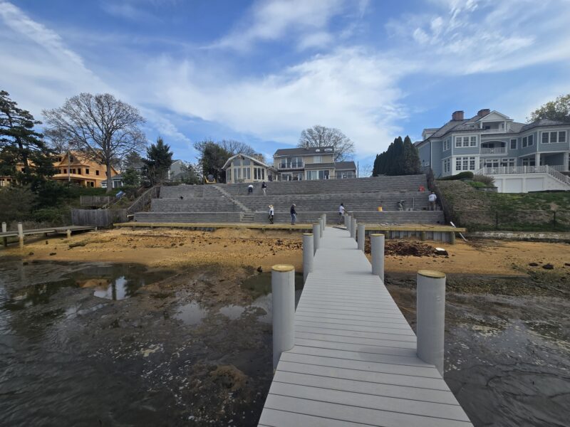 Long wooden boardwalk extends toward stone steps at a waterfront, with large houses on the hillside behind it.