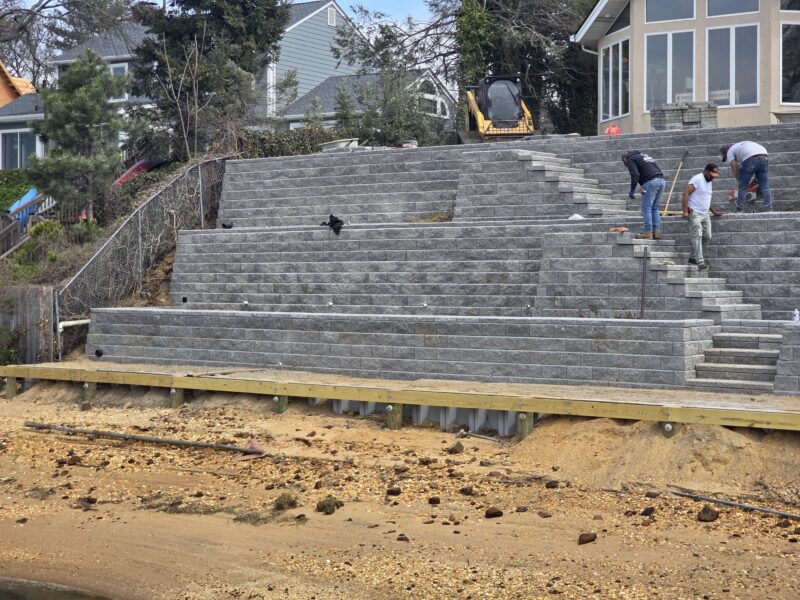 Construction workers building a masonry retaining wall on a hillside; a small excavator sits behind the wall.