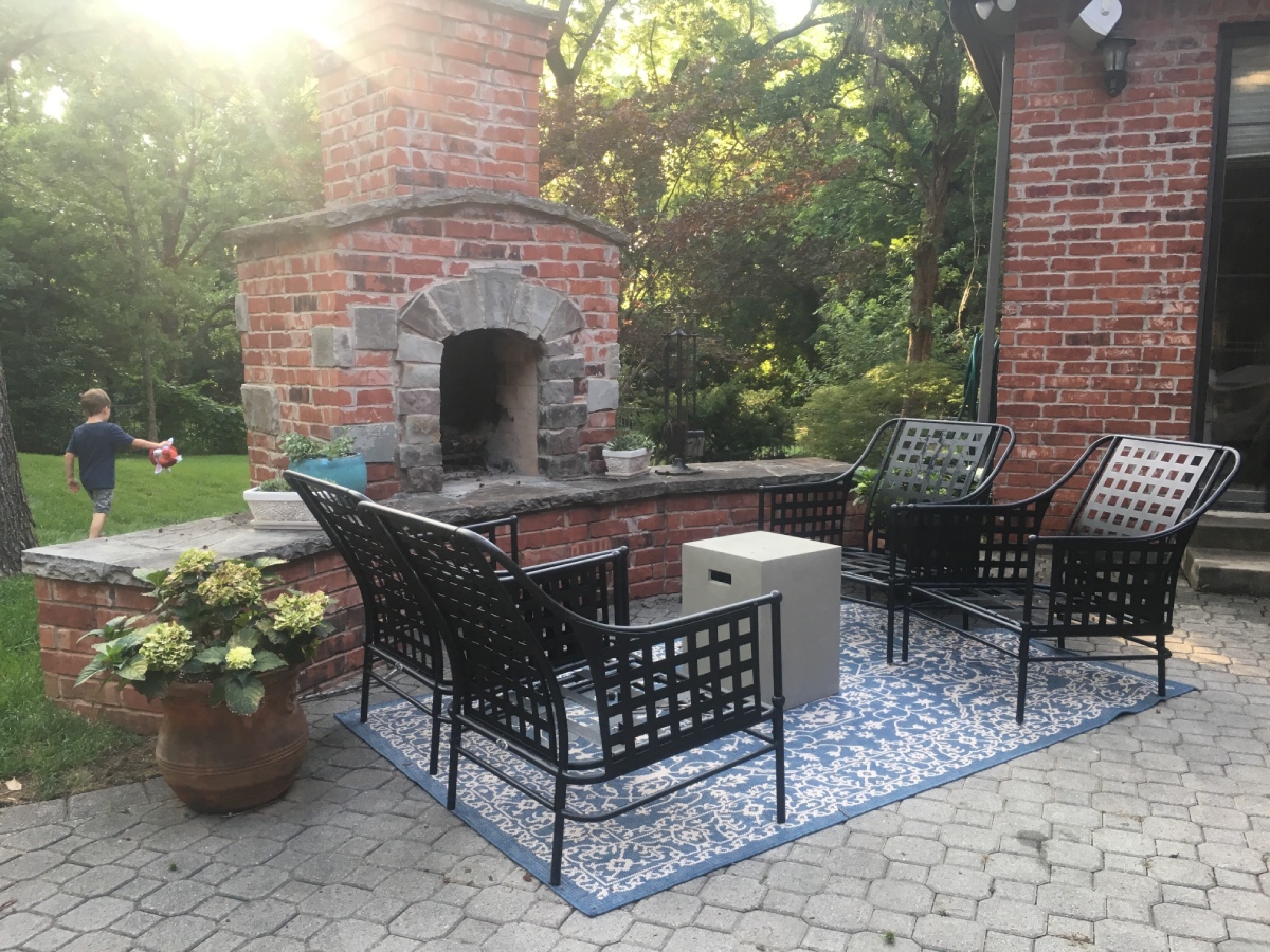 Outdoor brick patio with a stone fireplace, black metal seating around a blue patterned rug and a small square side table, potted plant nearby.
